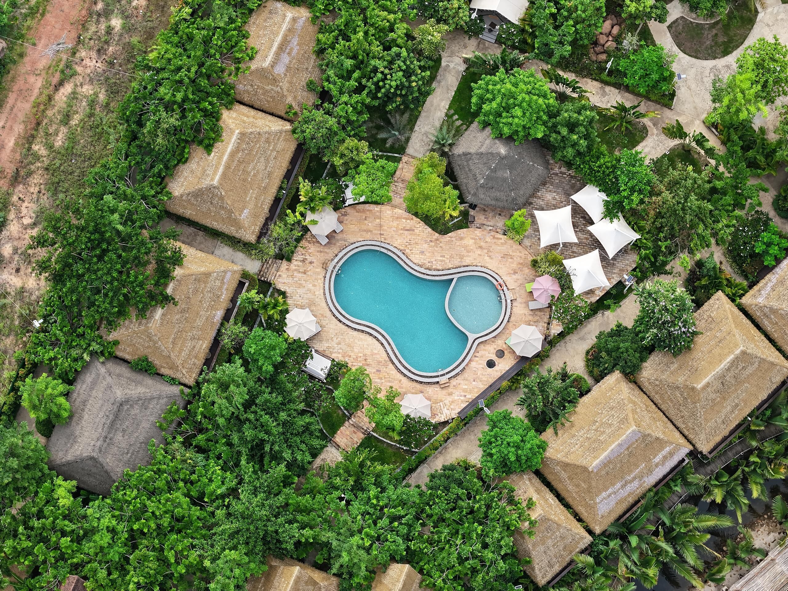 An aerial view of a resort with a heart-shaped pool surrounded by thatched-roof huts and lush greenery.