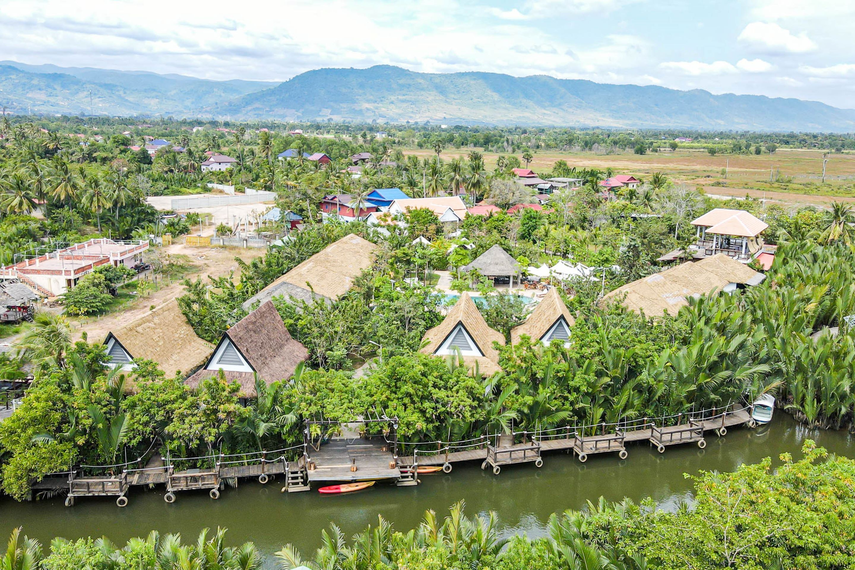 A resort with thatched-roof huts, a dock with canoes, and a river surrounded by lush greenery and mountains in the background.