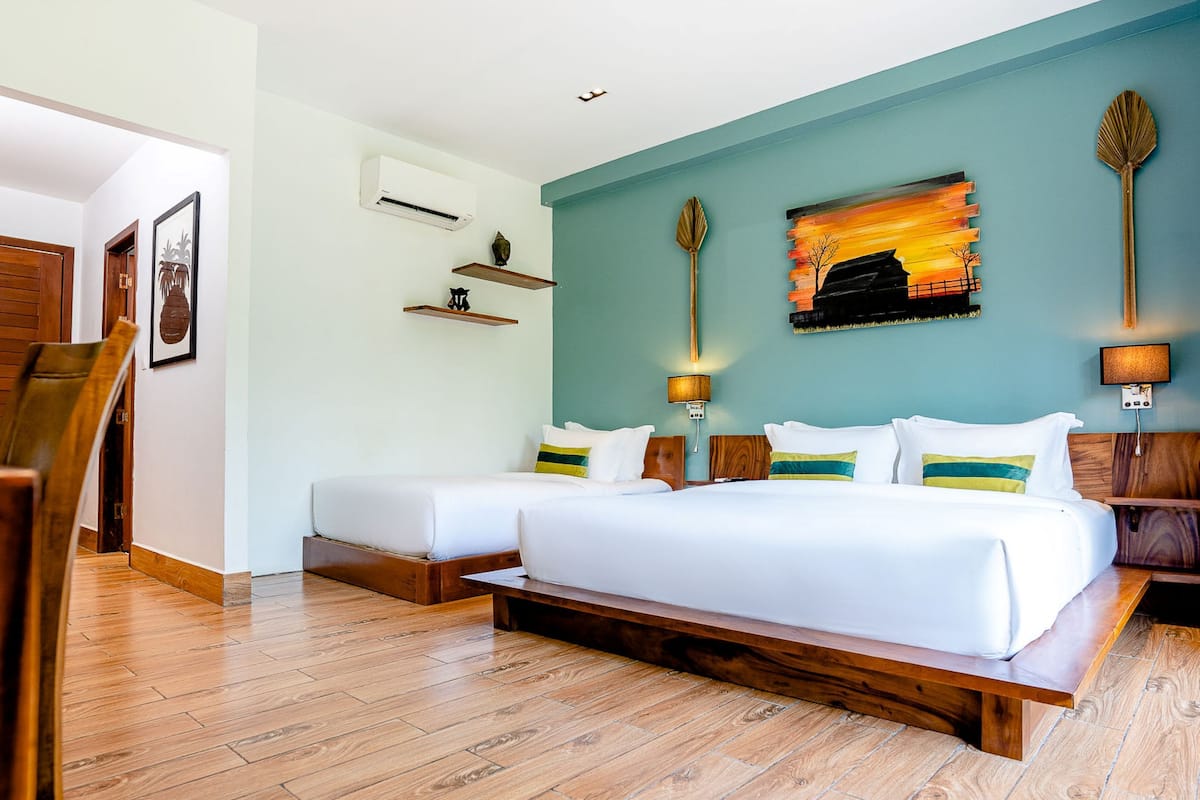 A hotel room with two beds, wooden flooring, a wall-mounted air conditioner, and a painting of a sunset.