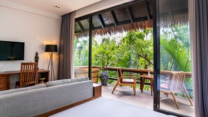 Habitación de hotel moderna con una cama grande, un escritorio, un televisor y un balcón con mobiliario de madera y vistas a exuberante vegetación.