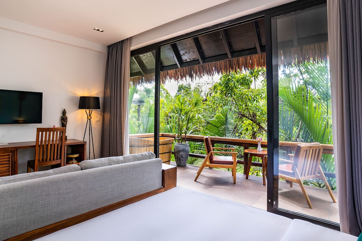 A modern hotel room with a large bed, a desk, a TV, and a balcony with wooden furniture and a view of lush greenery.