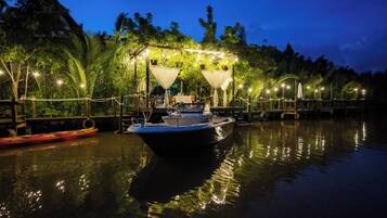 A boat docked at a pier with a canopy and string lights, surrounded by palm trees and a calm body of water.