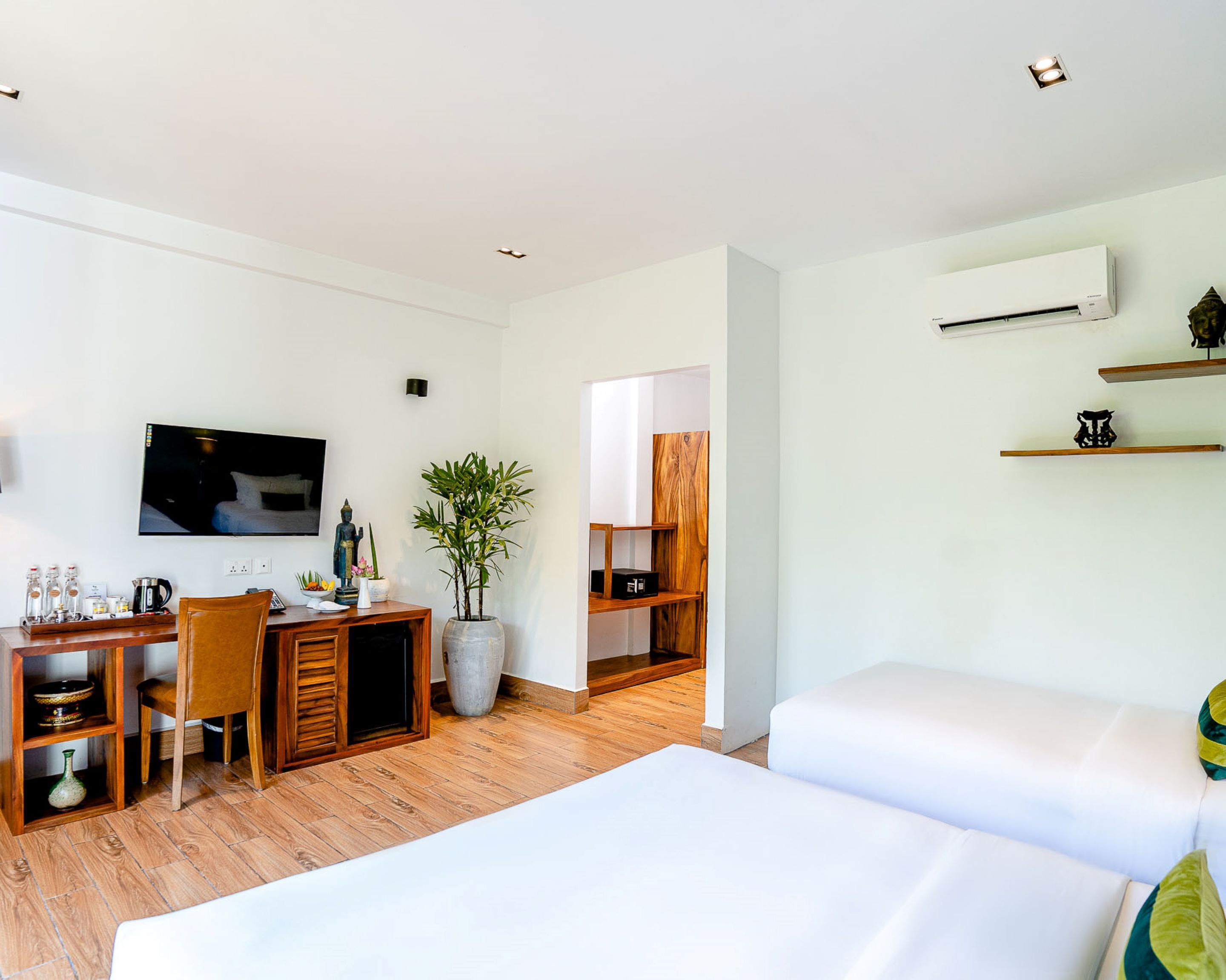 A modern hotel room with a wooden desk, a television, a large potted plant, and a bed with white bedding.