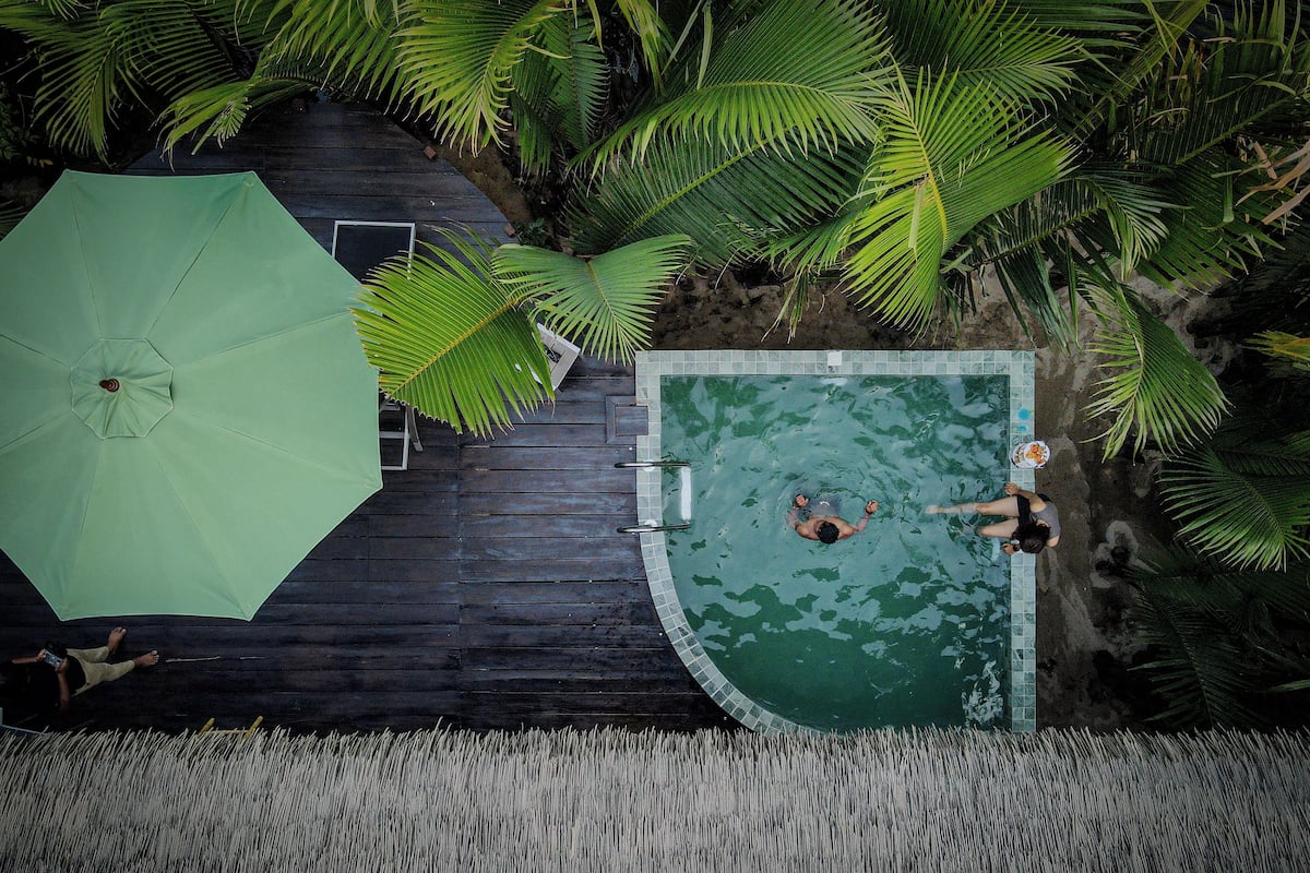 An aerial view of a swimming pool surrounded by palm trees, with a green umbrella and a wooden deck.