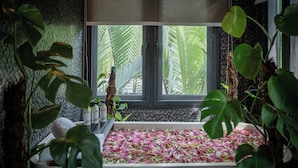 Una habitación con una bañera llena de pétalos de rosa, rodeada de exuberantes plantas verdes y una ventana con vista a palmeras.