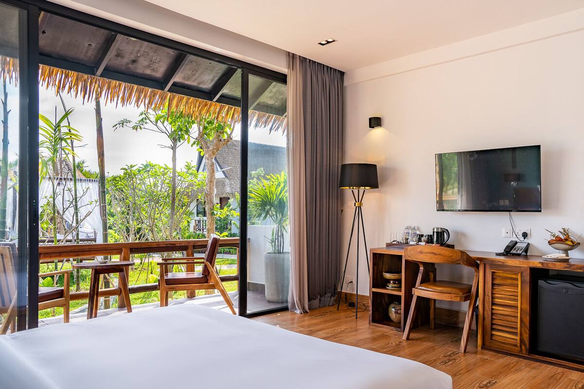 A hotel room with a large bed, a desk with a chair, a TV mounted on the wall, and a balcony with a view of greenery and a thatched roof structure.