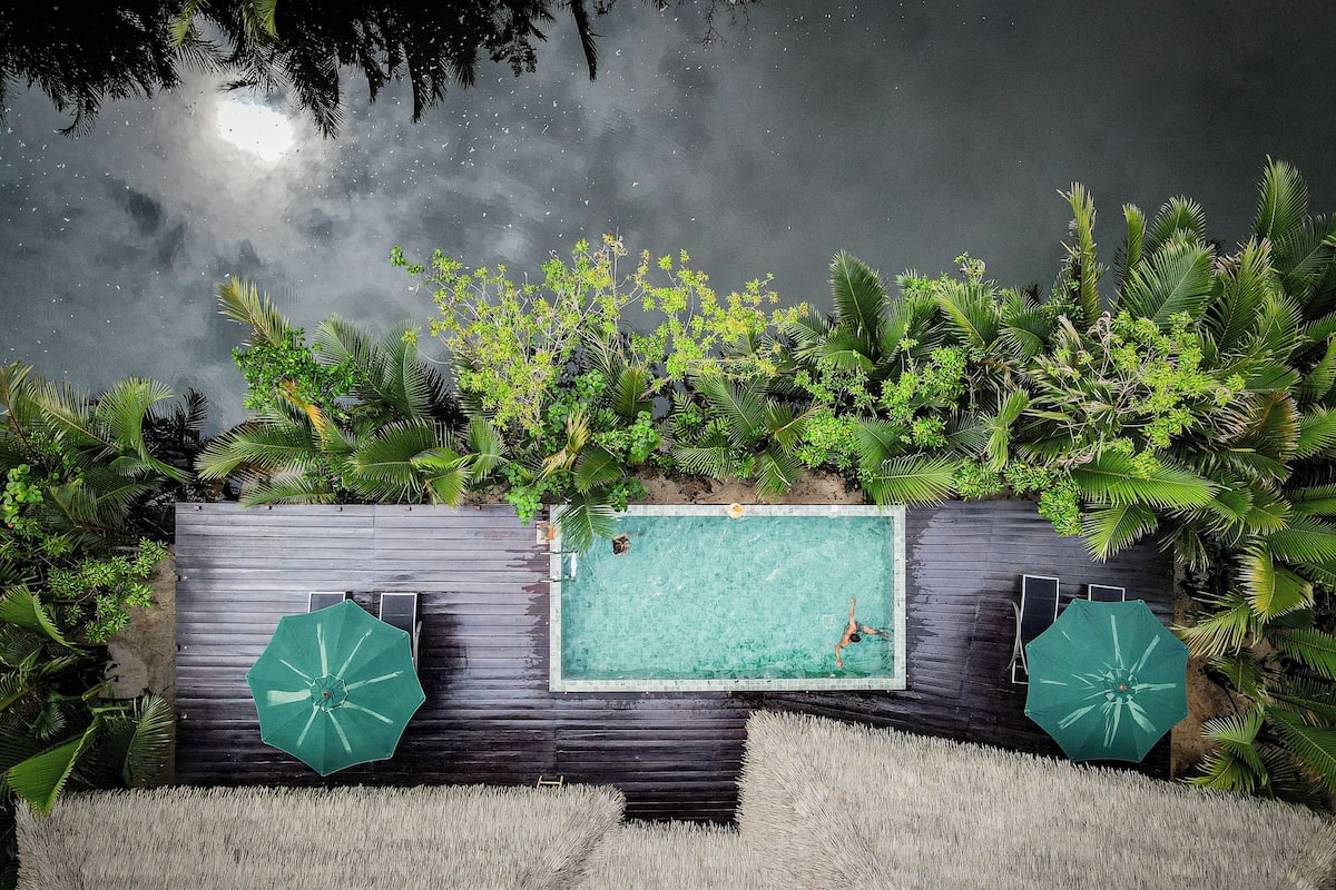 A rectangular swimming pool with two green umbrellas, surrounded by lush greenery and a wooden deck.