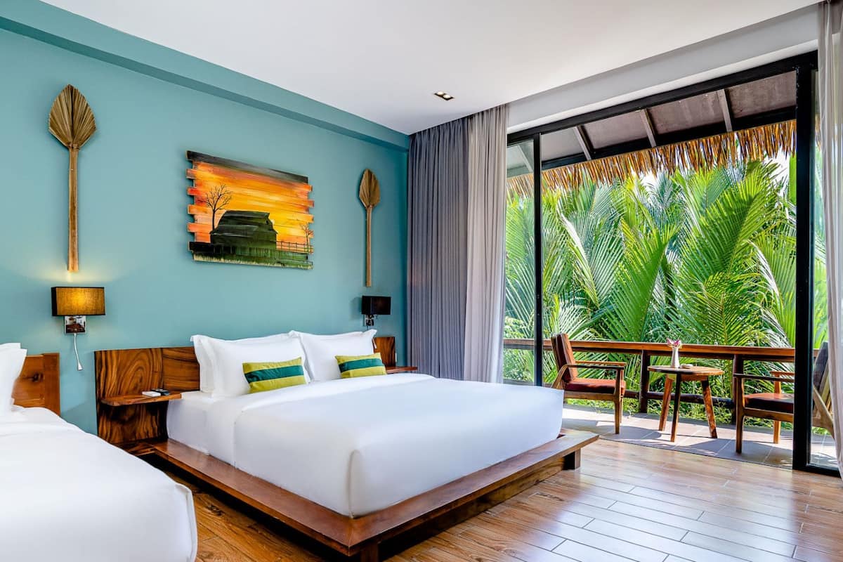 A hotel room with a large bed, a painting on the wall, and a view of palm trees through the balcony.