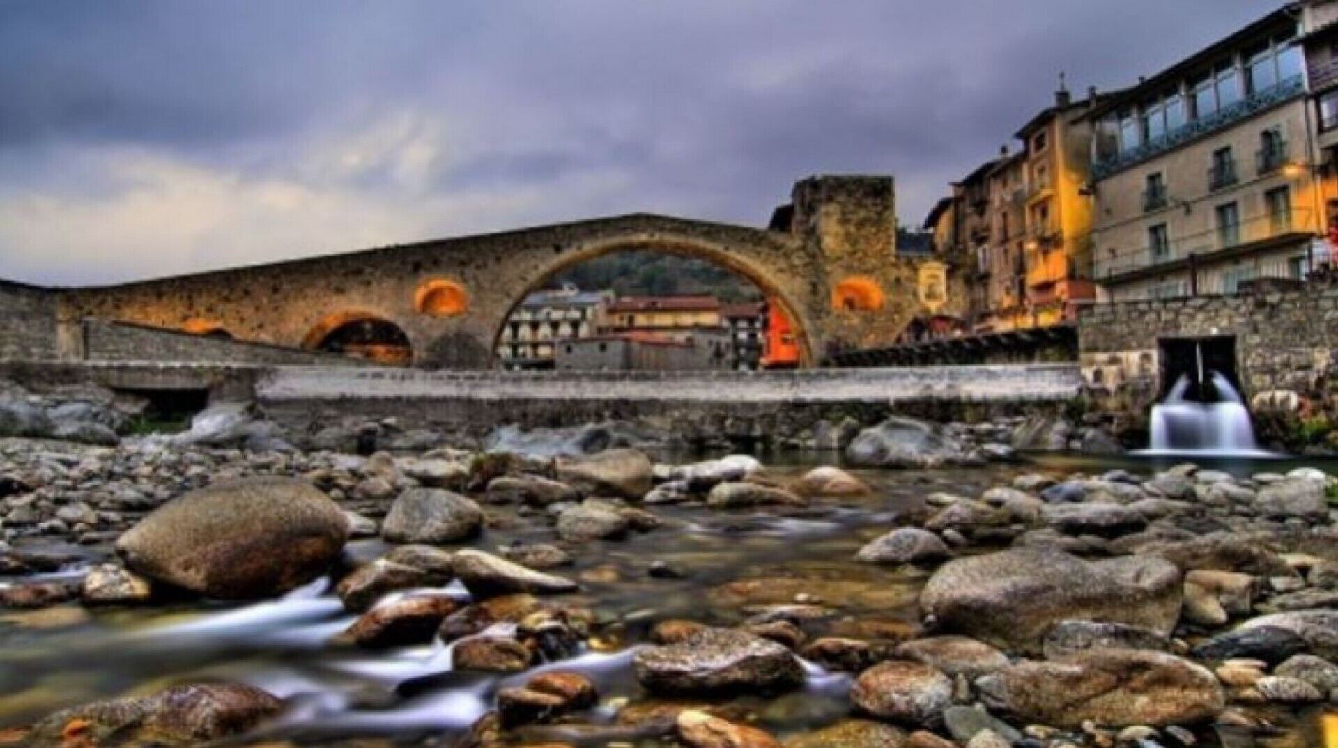 A stone bridge over a river with a waterfall, surrounded by rocky terrain and historic buildings.