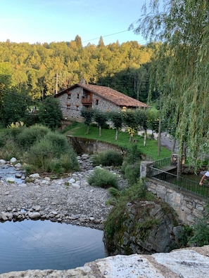 Una casa de piedra con balcón, rodeada de vegetación y un arroyo pequeño.