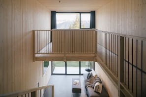 Habitación espaciosa con techos altos, interior de madera, balcón con barandilla, sofá y ventana con vistas al exterior.