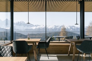 Un comedor moderno con mesas y sillas de madera, ventanas amplias con vistas a la montaña e iluminación colgante.