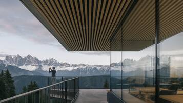 A balcony with a view of snow-capped mountains, a person taking a photo, and modern glass railings.