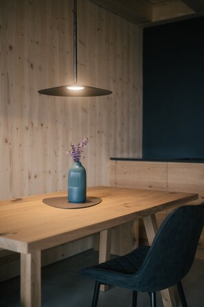 Une table de salle à manger en bois, ornée d’un vase de fleurs, un luminaire suspendu moderne et une chaise de couleur sombre.