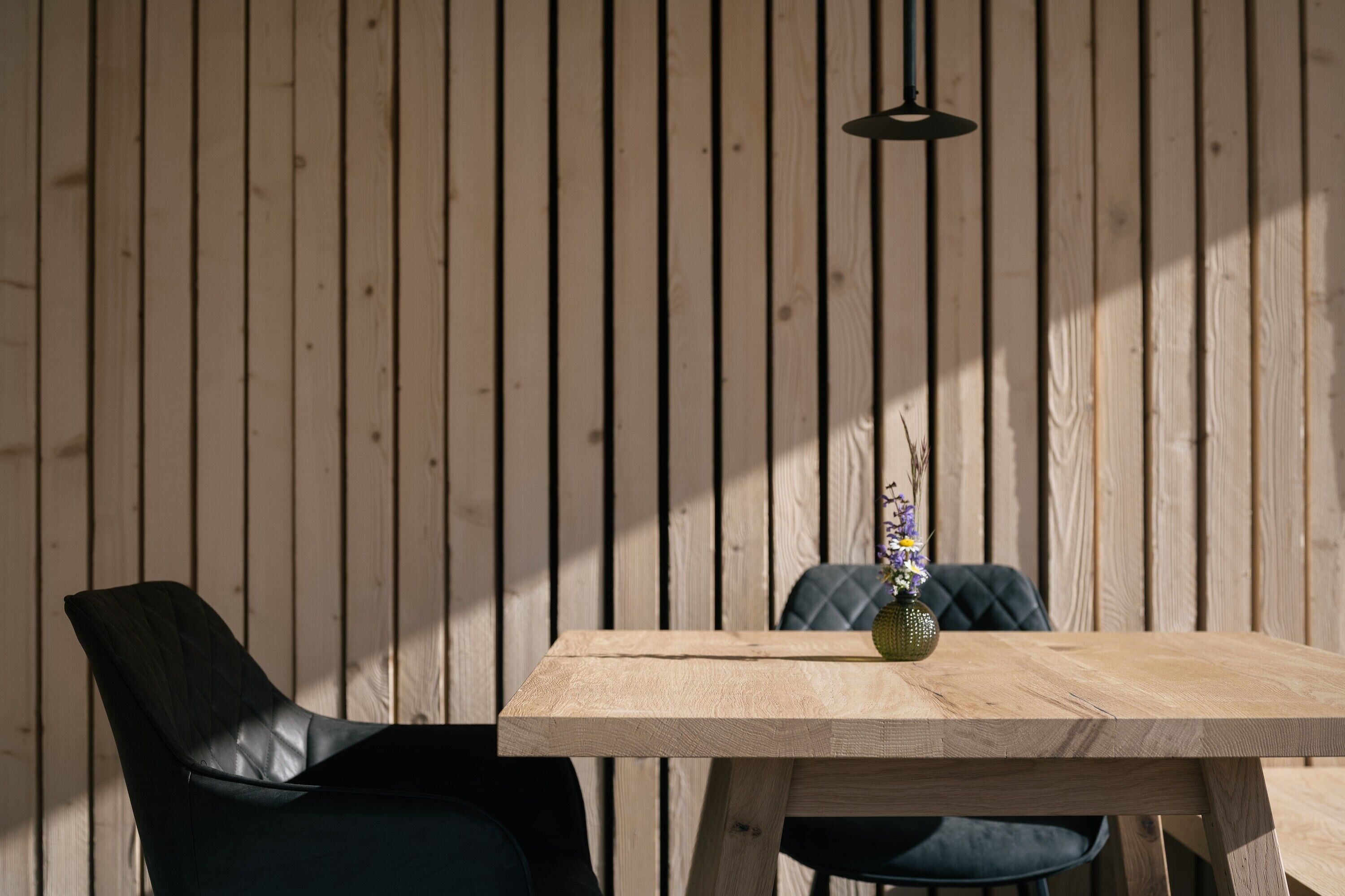 A wooden dining table with a vase of flowers, two chairs, and a hanging light fixture against a wooden wall.