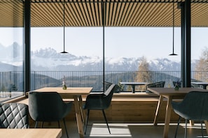 Un espace de restauration moderne avec des tables et des chaises en bois, de grandes fenêtres offrant une vue sur les montagnes et des luminaires suspendus.