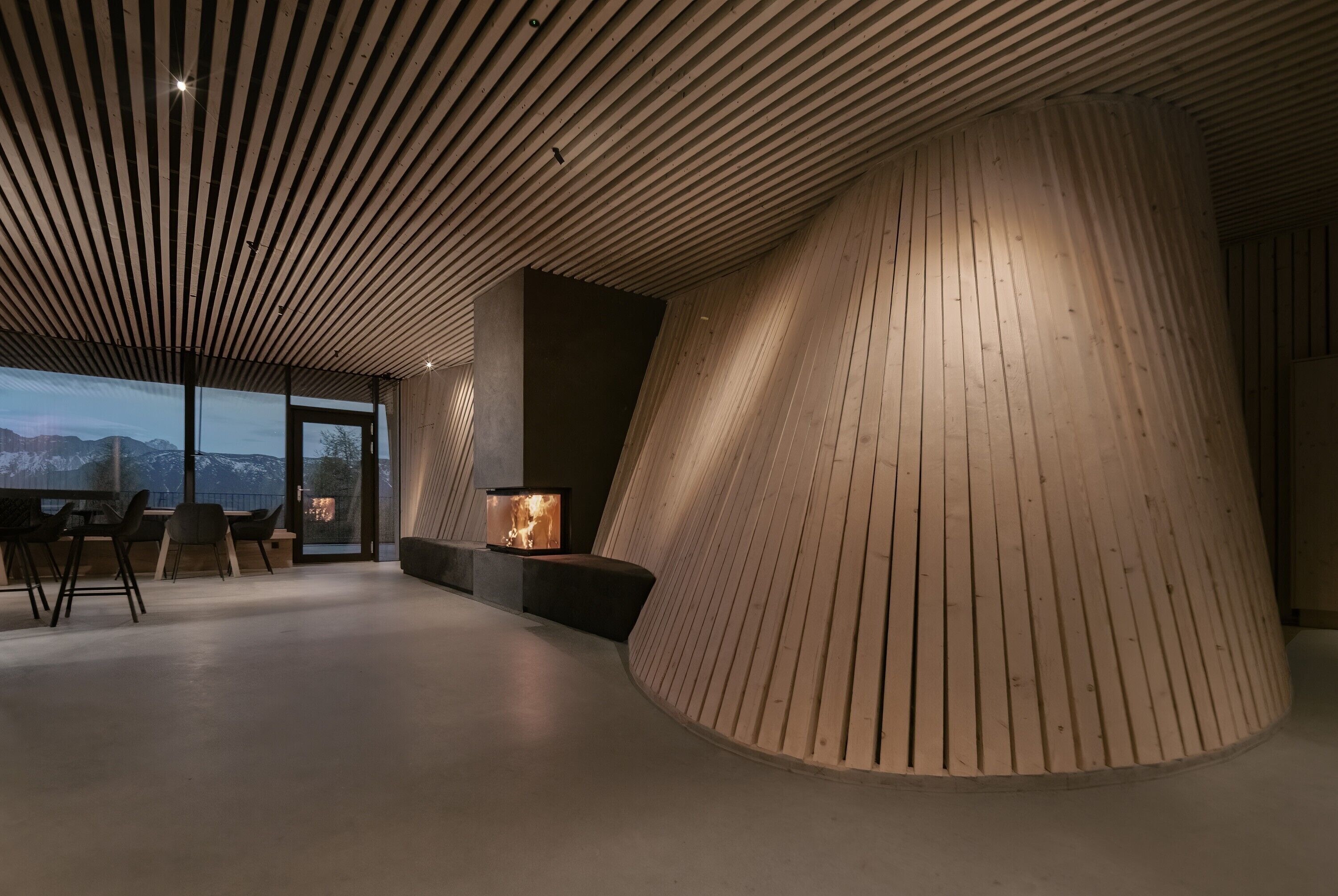 A modern interior with a curved wooden wall, a fireplace, and a dining area with a view of mountains.