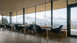A modern dining area with tables and chairs, large windows with mountain views, and a wooden ceiling.