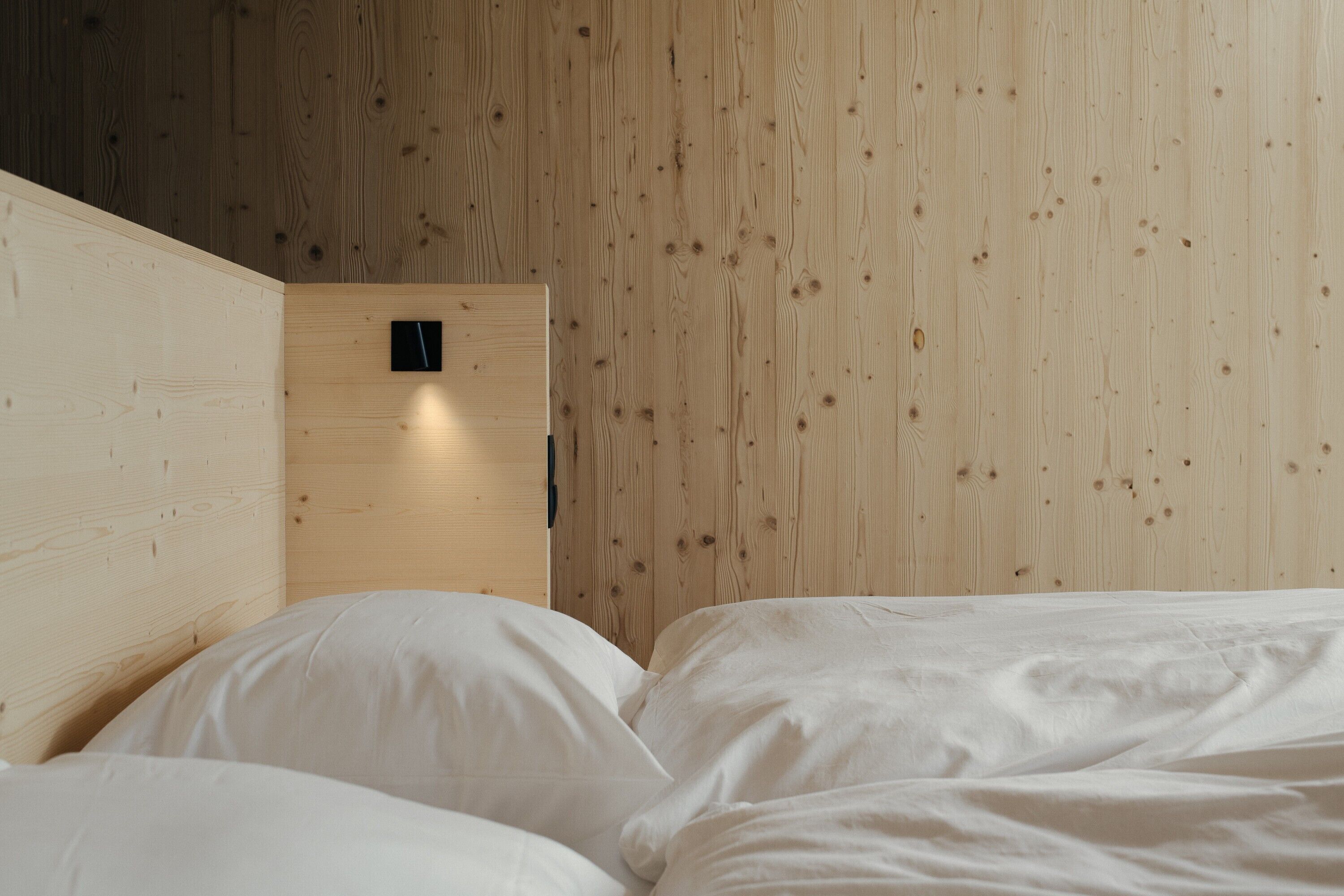 A room with a wooden wall, a bed with white bedding, and a small black object on the wall.