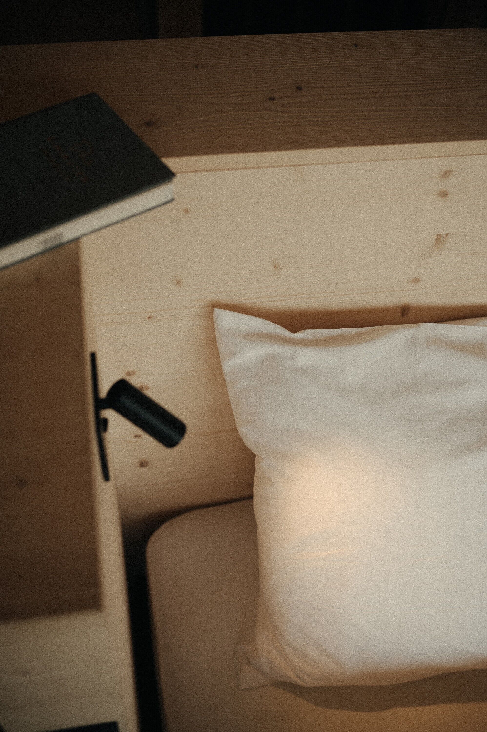 Ein hölzerner Kopfteil, ein weißes Kopfkissen, ein schwarzes Buch und eine schwarze Lampe auf einem Nachttisch aus Holz.