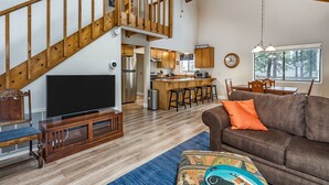 A spacious living area with a wooden staircase, a kitchen with bar stools, a large flat-screen TV, and a cozy seating area with a patterned ottoman. - Three Wild Turkeys - Amazing Outdoor Spaces!  2 Decks! by Yosemite Region Resorts (Groveland)