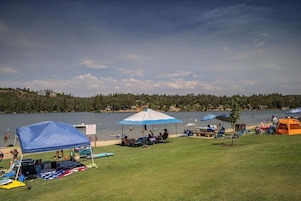 A lakeside camping area with tents, a covered seating area, and a grassy field.