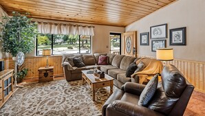 A cozy living room with wooden ceiling and walls, a large window with curtains, a sofa, a coffee table, and a lamp. - Bigfoot Retreat - Close to Golf and the Beach by Yosemite Region Resorts (Groveland)
