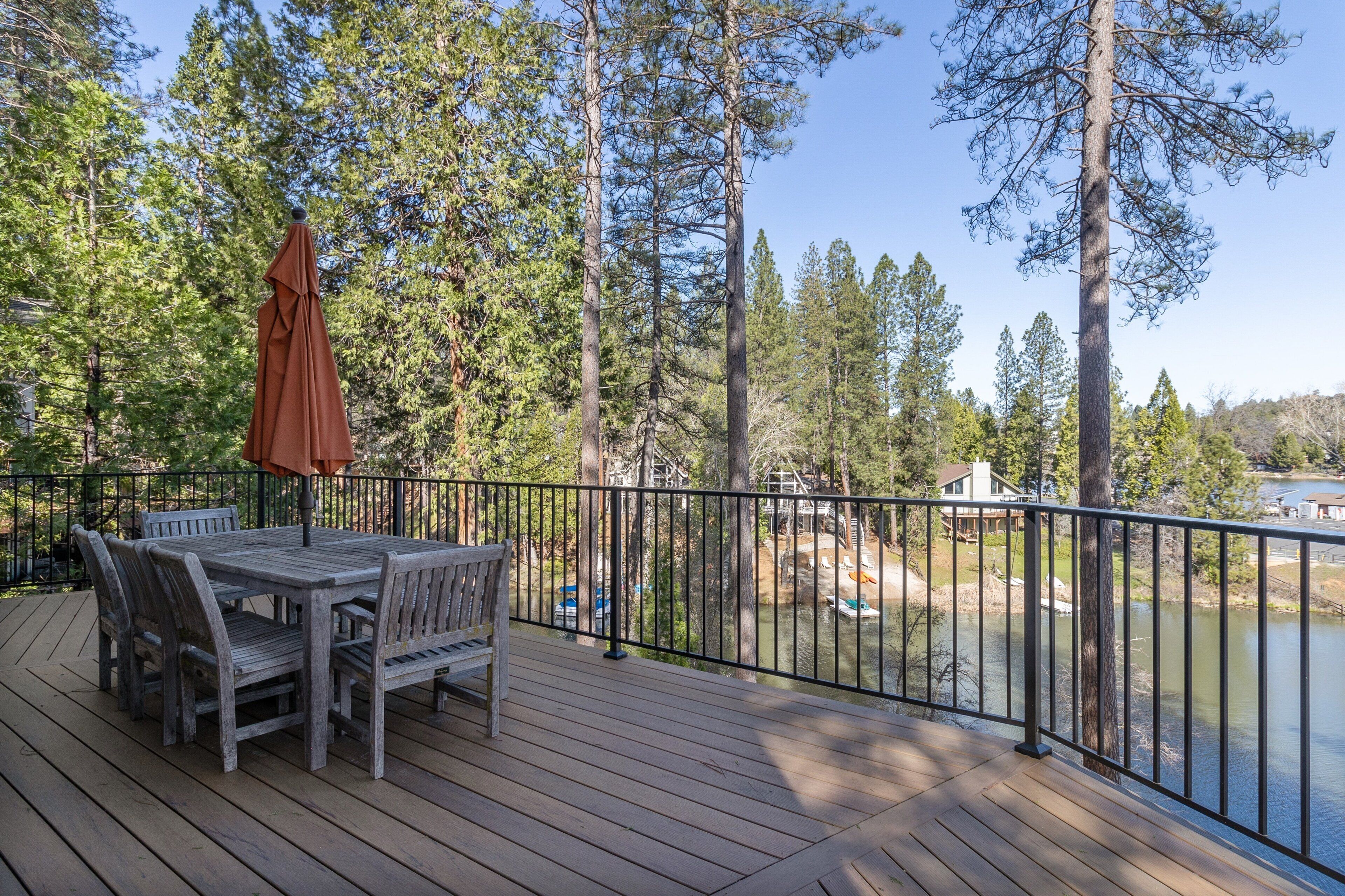 A wooden deck with a table and chairs, overlooking a lake with a view of trees and a distant building.