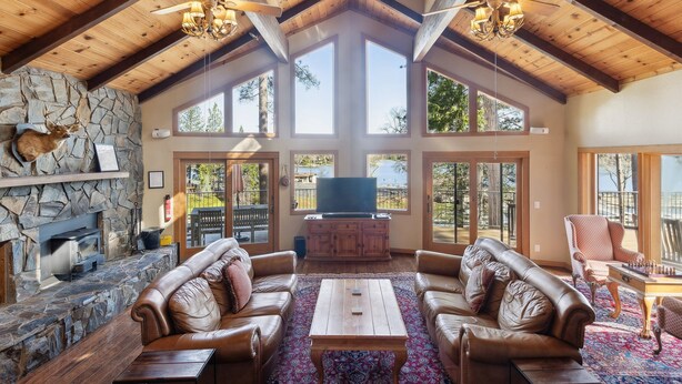 A spacious living room with leather sofas, a wooden coffee table, and a stone fireplace. There are large windows and a ceiling fan. - Dos Pinos - Spacious Lakefront Cabin w/ Game Room by Yosemite Region Resorts (Groveland)