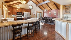 A spacious kitchen with wooden beams, a large island with bar stools, and a dining area with a table and chairs. - Dos Pinos - Spacious Lakefront Cabin w/ Game Room by Yosemite Region Resorts (Groveland)