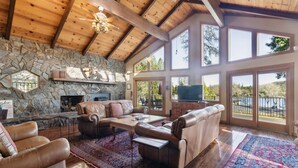 A spacious living room with a stone fireplace, leather furniture, and large windows overlooking a scenic view. - Dos Pinos - Spacious Lakefront Cabin w/ Game Room by Yosemite Region Resorts (Groveland)