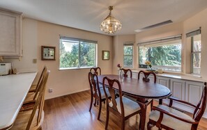 A dining room with a round table, wooden chairs, and a chandelier. There are large windows with blinds, and a kitchen area is visible to the left. - Casa Del Lago - Only 200 Yards from the Beach by Yosemite Region Resorts (Groveland)
