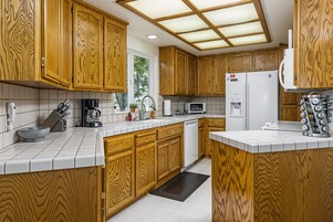 A kitchen with wooden cabinets, a white countertop, a microwave, a refrigerator, and a window with a view of trees.
