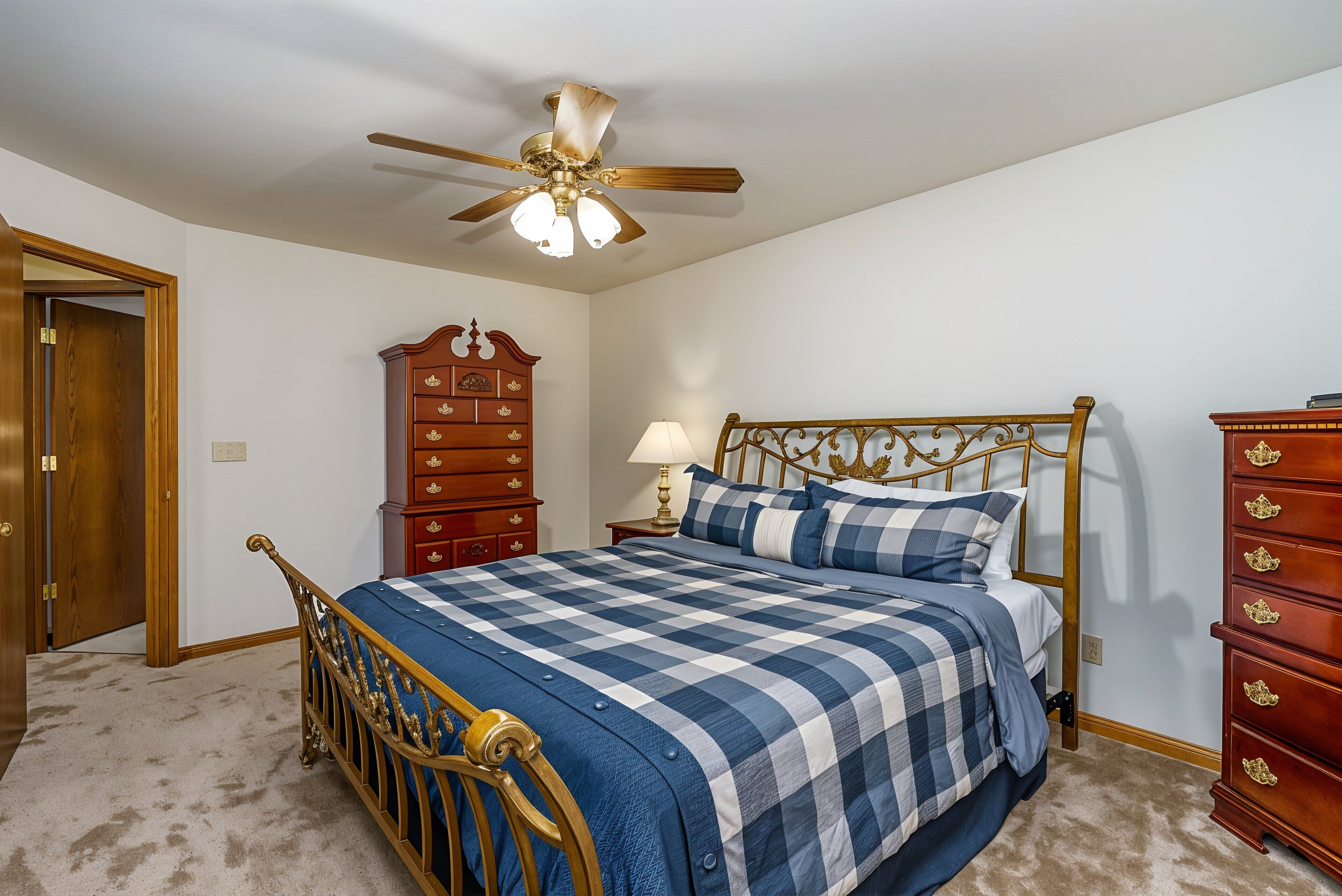 A bedroom with a wooden bed frame, a checkered bedspread, a nightstand with a lamp, and a dresser with drawers.