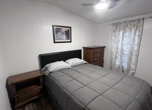 A bedroom with a bed, a nightstand, a dresser, a framed picture on the wall, and a window with curtains.