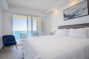 Relax in this serene hotel room with a large bed, a blue armchair, and a side table with a lamp, offering a stunning sea view.