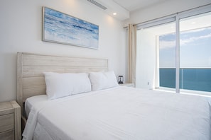 Une chambre avec un grand lit, une table de chevet, une commode, un tableau et une vue sur la mer, visible à travers une porte coulissante.