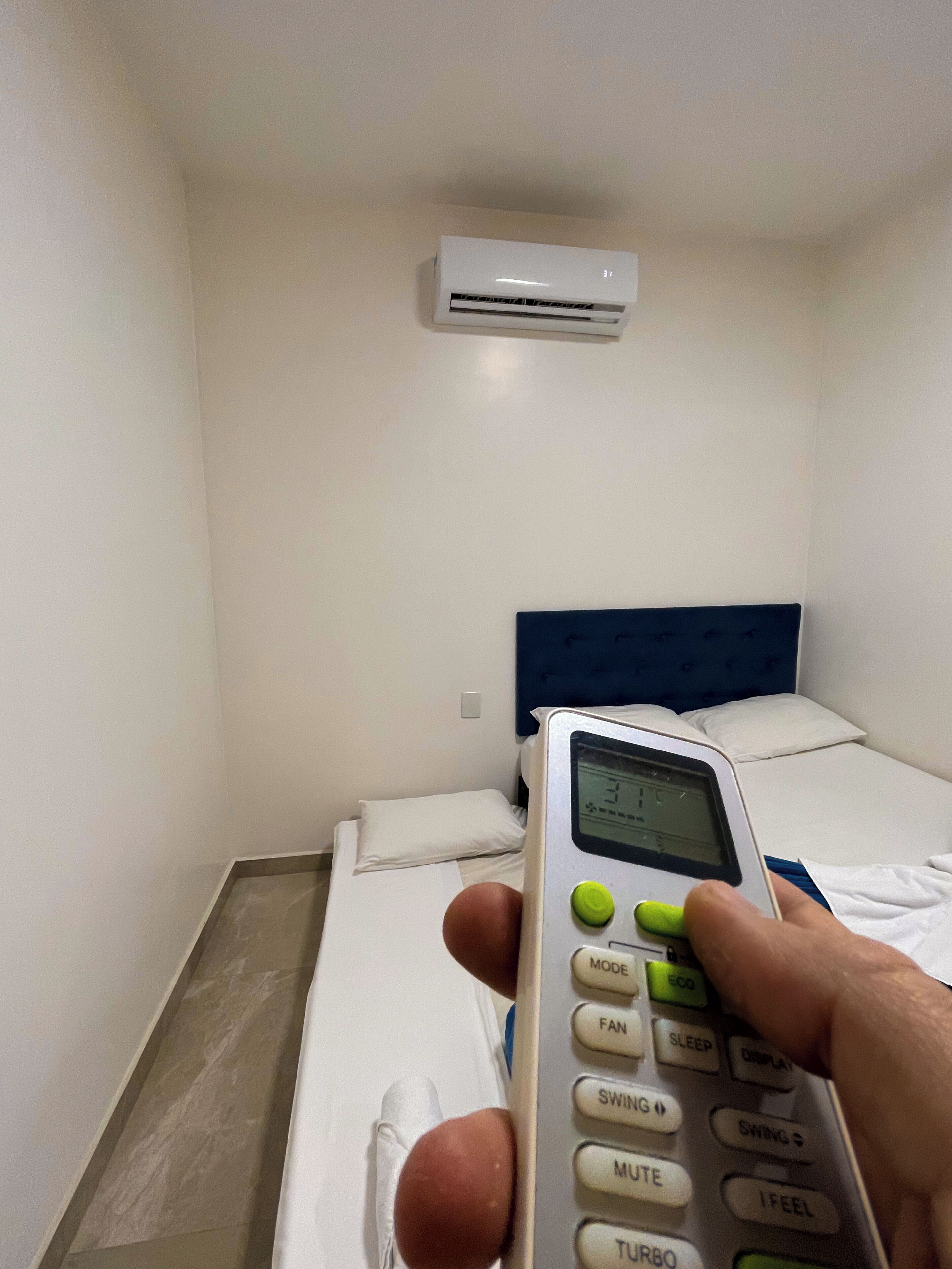 A hotel room with a bed, a bedside table, an air conditioning unit, and a control panel.