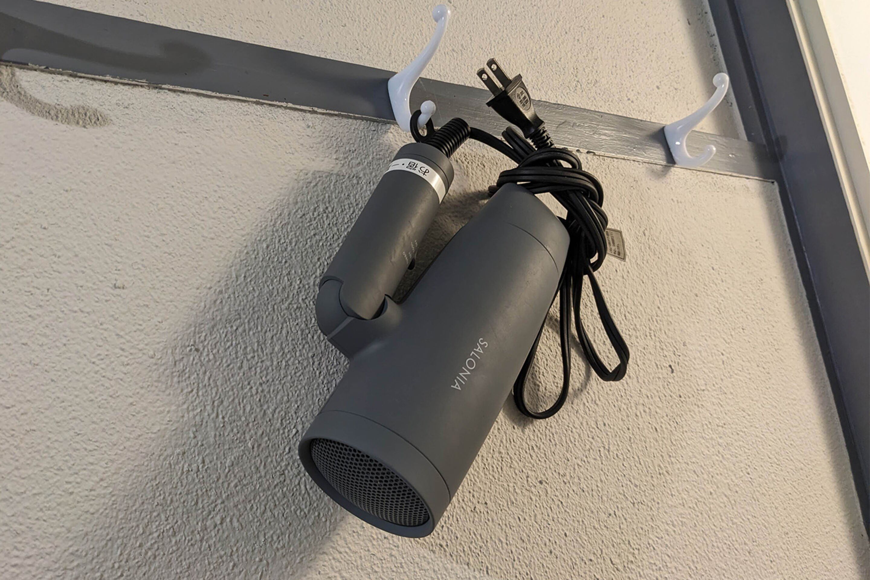 A black device with a cord and a plug, mounted on a textured wall with two white hooks.