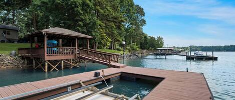 A wooden dock extending into a lake, with a covered seating area and a small pavilion.