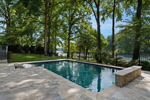 Une piscine rectangulaire avec une terrasse et un banc en pierre, entourée d’une clôture noire et d’arbres.