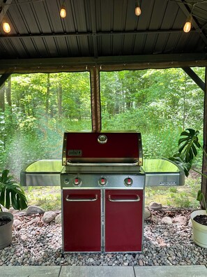 Un barbecue rouge pour lâextĂ©rieur, ornĂ© du logo Genesis, placĂ© sous un espace abritĂ© avec vue sur une forĂȘt.