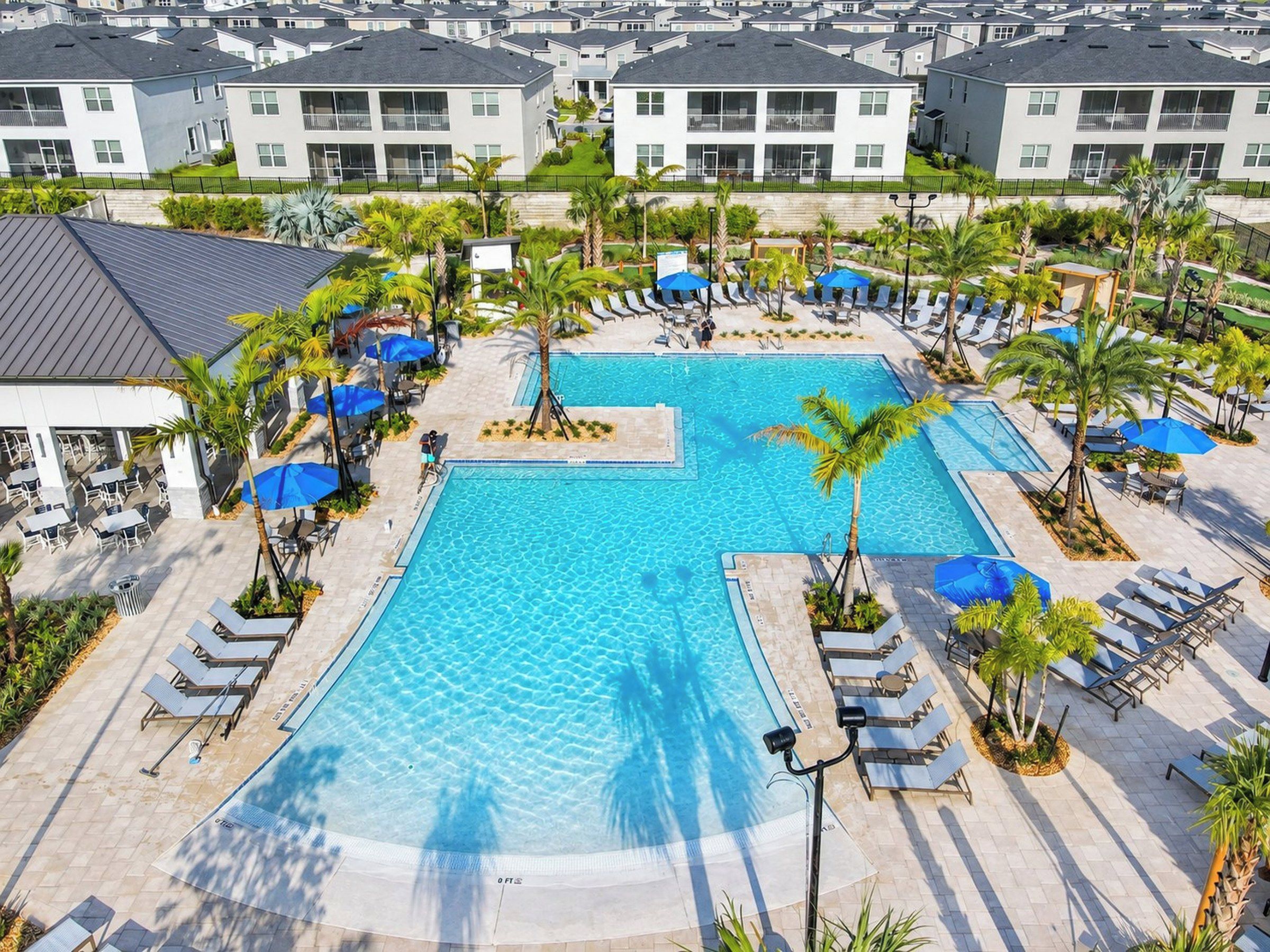 A large swimming pool surrounded by lounge chairs and umbrellas, with a view of apartment buildings in the background.