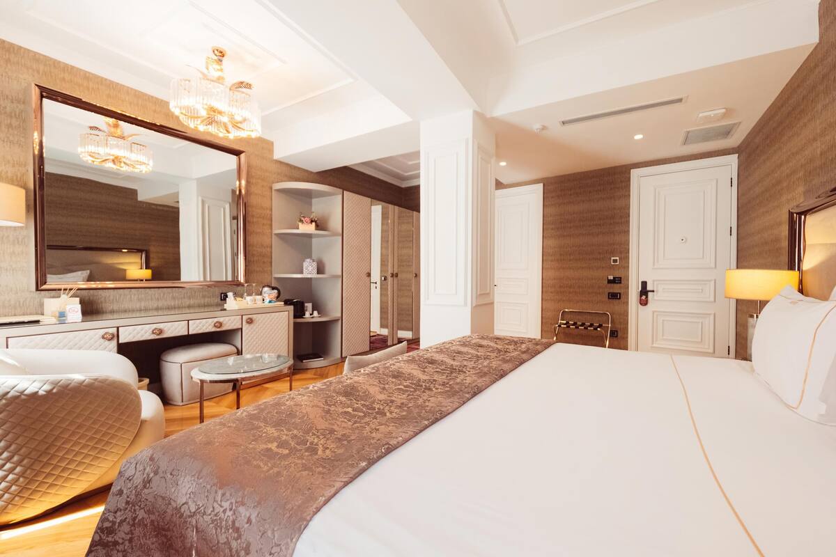 A modern hotel room with a large bed, a vanity with a mirror, a chandelier, and a view of the city.