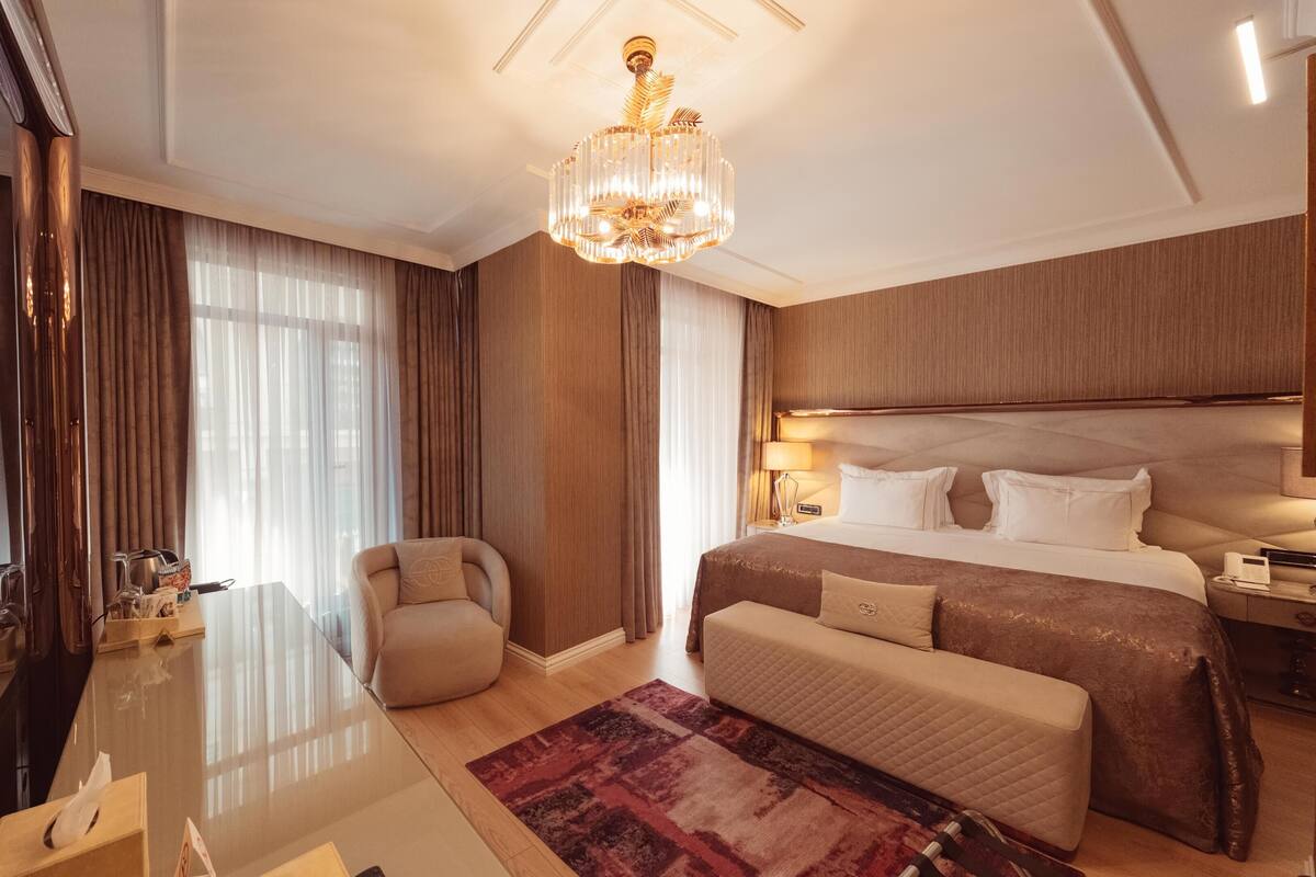 A hotel room with a large bed, a desk, a chair, a chandelier, and a view of the outdoors.