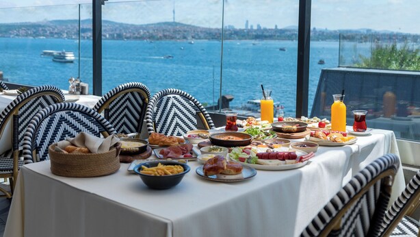 Una mesa dispuesta con diferentes platos y bebidas, con vistas a un cuerpo de agua con barcos y un paisaje urbano lejano.