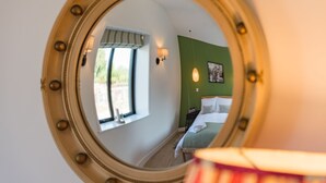 Ein runder Spiegel mit dekorativem Rahmen, der einen Raum mit einem Bett, einem Fenster und einer Wandlampe reflektiert.