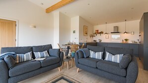 Ein modernes Wohnzimmer mit zwei grauen Sofas, einem Holzbalken an der Decke, einem Essbereich und einer Küche mit Pendelleuchten.
