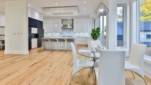 Uma cozinha moderna com armários brancos, uma ilha central com banquetas e uma área de refeições com mesa de vidro e cadeiras brancas.