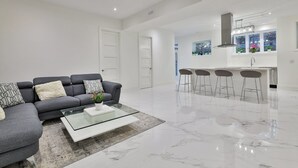 A modern living room with a grey sofa, a glass coffee table, and a kitchen area with a bar and stools.